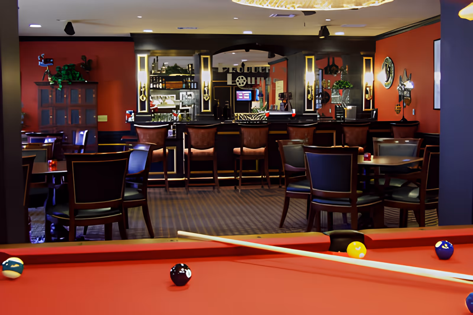 Interior view of a senior living facility lounge area featuring a red pool table with billiard balls and a cue stick in the foreground. Behind the pool table, there are several tables and chairs arranged for seating, and a bar area with high stools and decorative lighting. The walls are painted red with various decorations and mirrors.