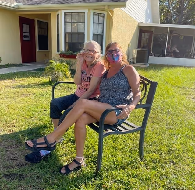 Two women sitting on a metal bench outside on a grassy lawn in front of a yellow building. One woman is wearing a pink shirt and glasses, smiling, and the other woman is wearing a patterned sleeveless top and a colorful face mask. The background shows windows, a door, and a screened porch area.