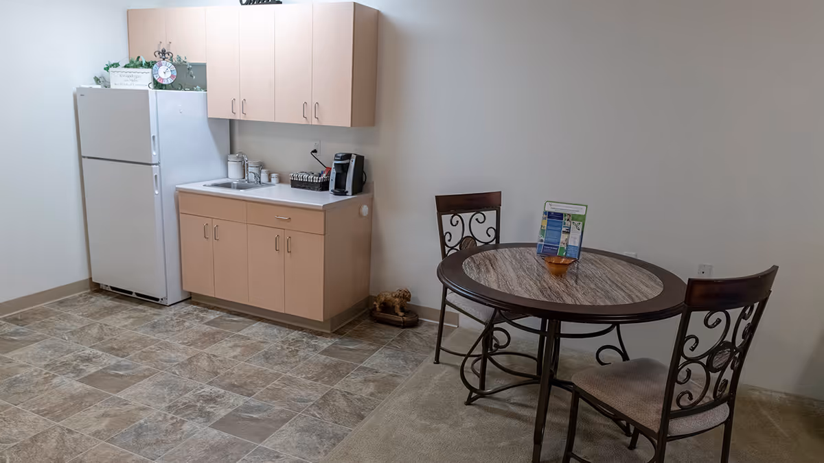 A small kitchenette area with light wood cabinets, a white refrigerator, a sink, and a coffee maker on the counter. Adjacent to the kitchenette is a round table with a marble-like top and two chairs with decorative backs. The floor is tiled near the kitchenette and carpeted near the table. A small decorative dog figurine is on the floor near the wall.