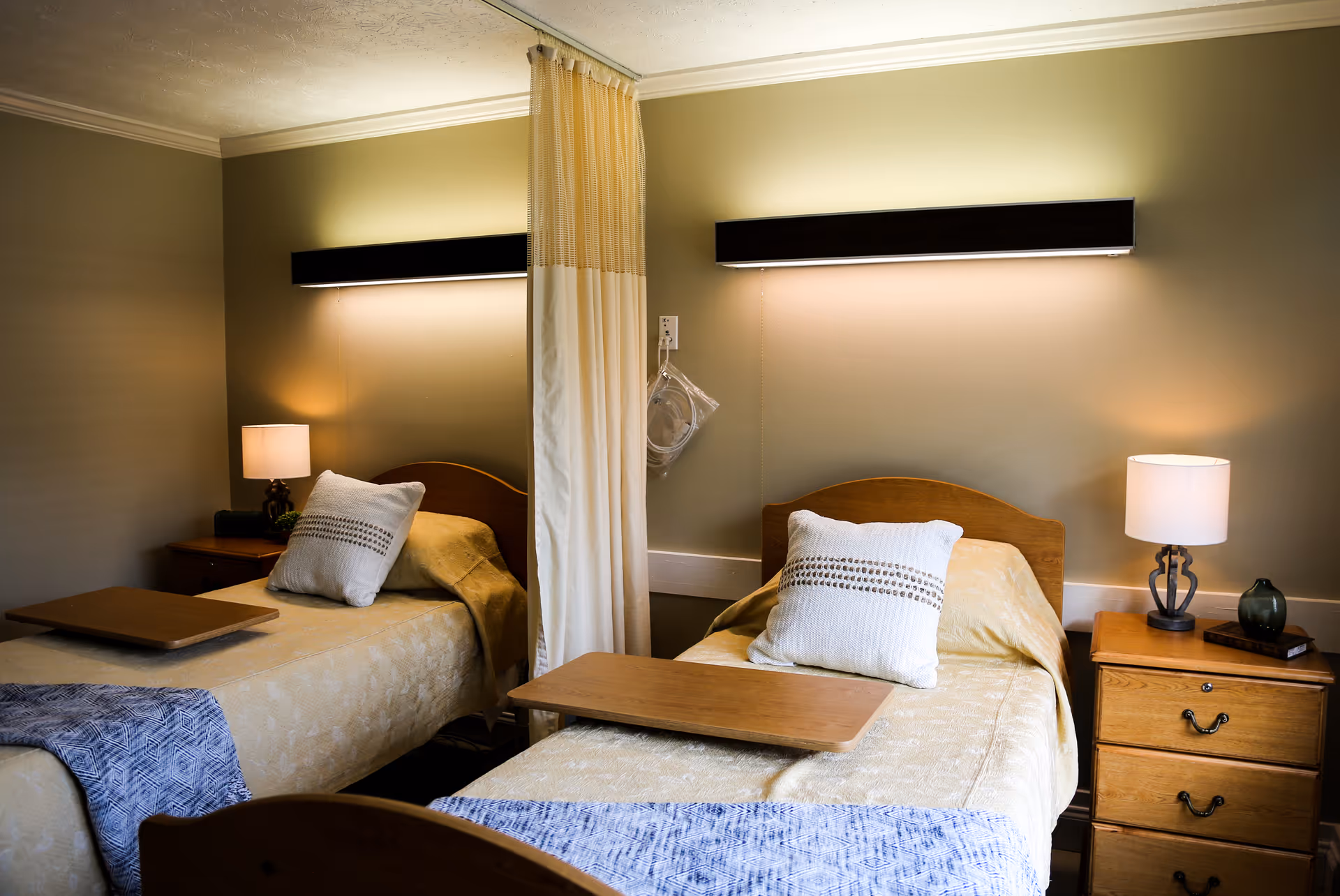 A shared bedroom in a senior living facility with two single beds separated by a privacy curtain. Each bed has a pillow and a beige bedspread, with a wooden overbed table. There are nightstands with lamps on either side of the beds, and soft wall-mounted lights above each bed.