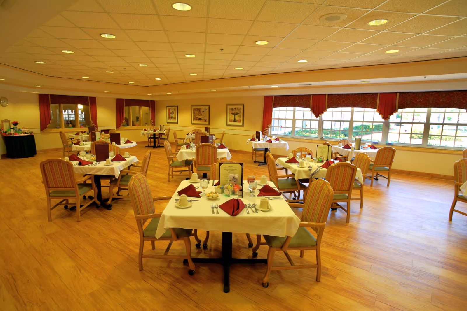 A spacious dining room with multiple tables covered in white tablecloths, each set with cups, glasses, silverware, and folded red napkins. The room has wooden flooring, large windows with red valances letting in natural light, and framed artwork on the walls. Chairs with striped upholstery surround the tables.