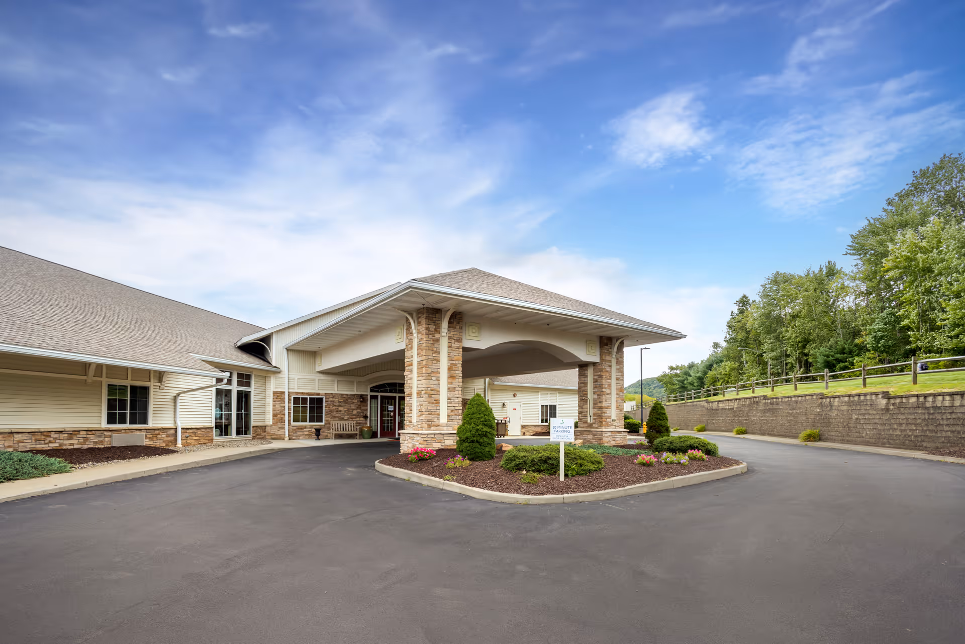 Front entrance of a one-story senior living facility with a covered porte-cochère, landscaped island, and paved driveway under a blue sky.