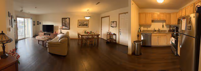 Panoramic view of a senior living apartment interior showing a living room with a beige sofa, coffee table, TV on a stand, and wall art. Adjacent to the living room is a dining area with a wooden dining table and chairs, and a kitchen with wooden cabinets, stainless steel refrigerator, stove, and a trash can on a dark wood floor.