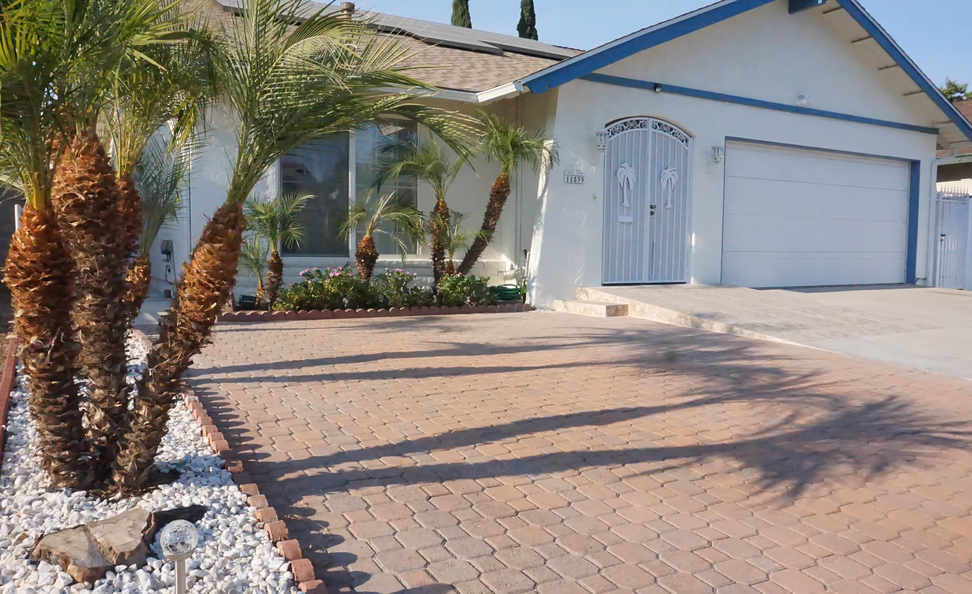 Front exterior view of a single-story house with a paved driveway, white walls, a white garage door, and a white decorative metal gate with palm tree designs. There are several palm trees and plants in a landscaped area with white rocks along the driveway.