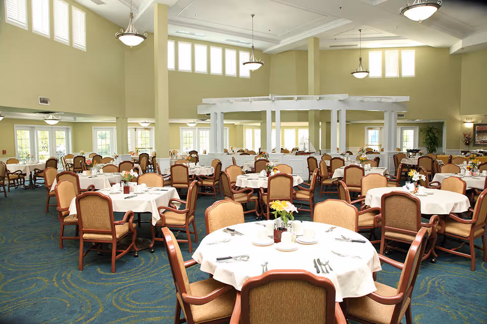A spacious dining room with multiple round tables covered with white tablecloths, each set with plates, cups, and silverware. The room has high ceilings with large windows allowing natural light to fill the space. Comfortable wooden chairs with cushioned seats surround each table. There are decorative flowers placed on each table, and the carpet has a blue and green pattern.