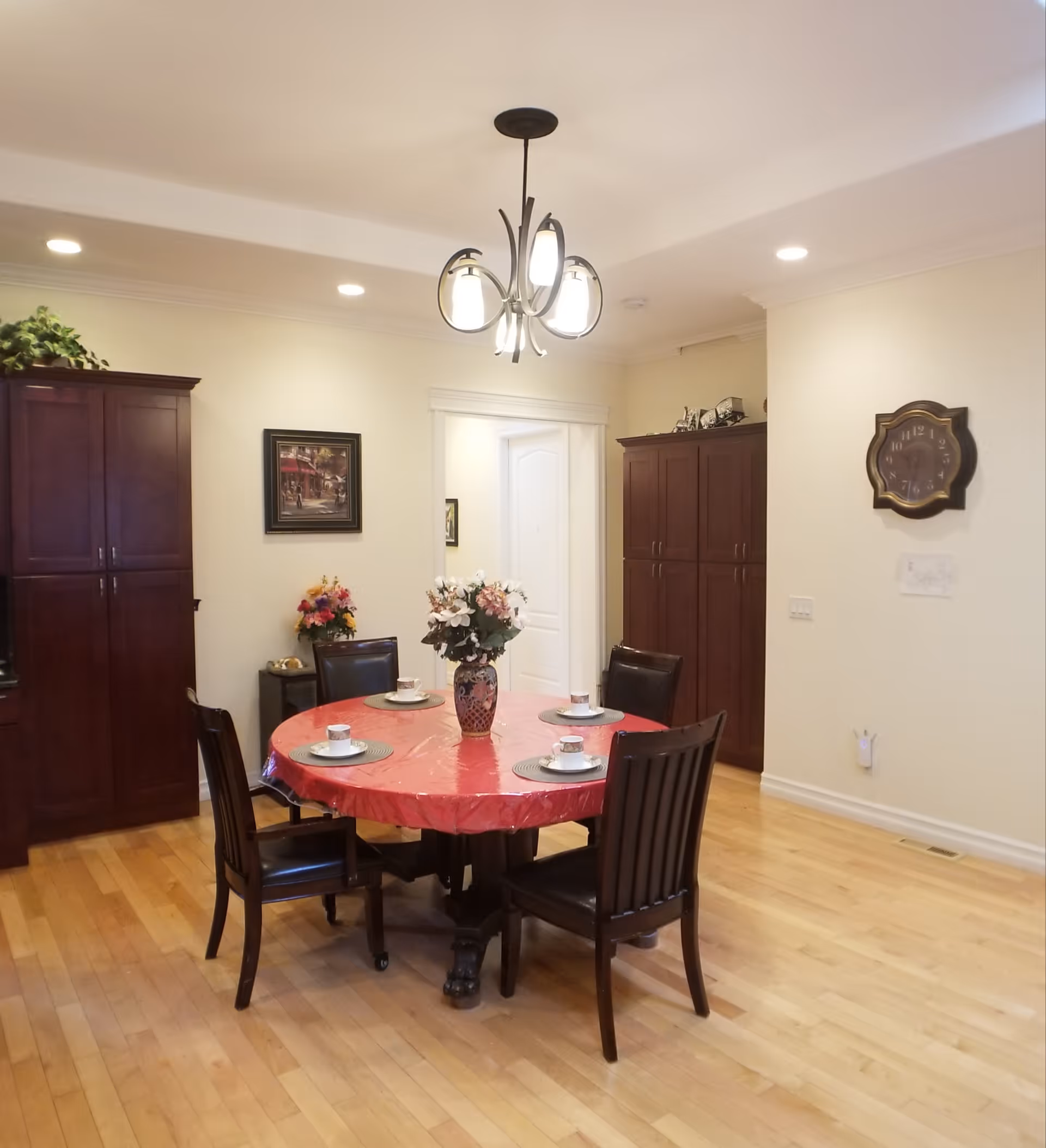 A dining room with a round table covered with a red tablecloth, set with four place settings including cups and saucers. The table has a floral centerpiece. Surrounding the table are four dark wooden chairs with black cushions. The room has light-colored walls, wooden flooring, two tall dark wooden cabinets, a wall clock, and a ceiling light fixture with three bulbs.