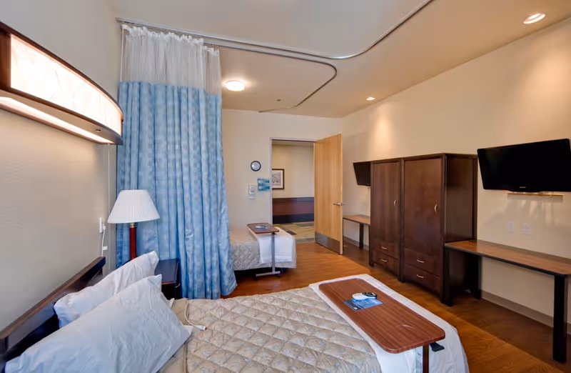 Shared senior living bedroom with two beds separated by a blue privacy curtain, wooden wardrobes and desks, and wall-mounted TVs.