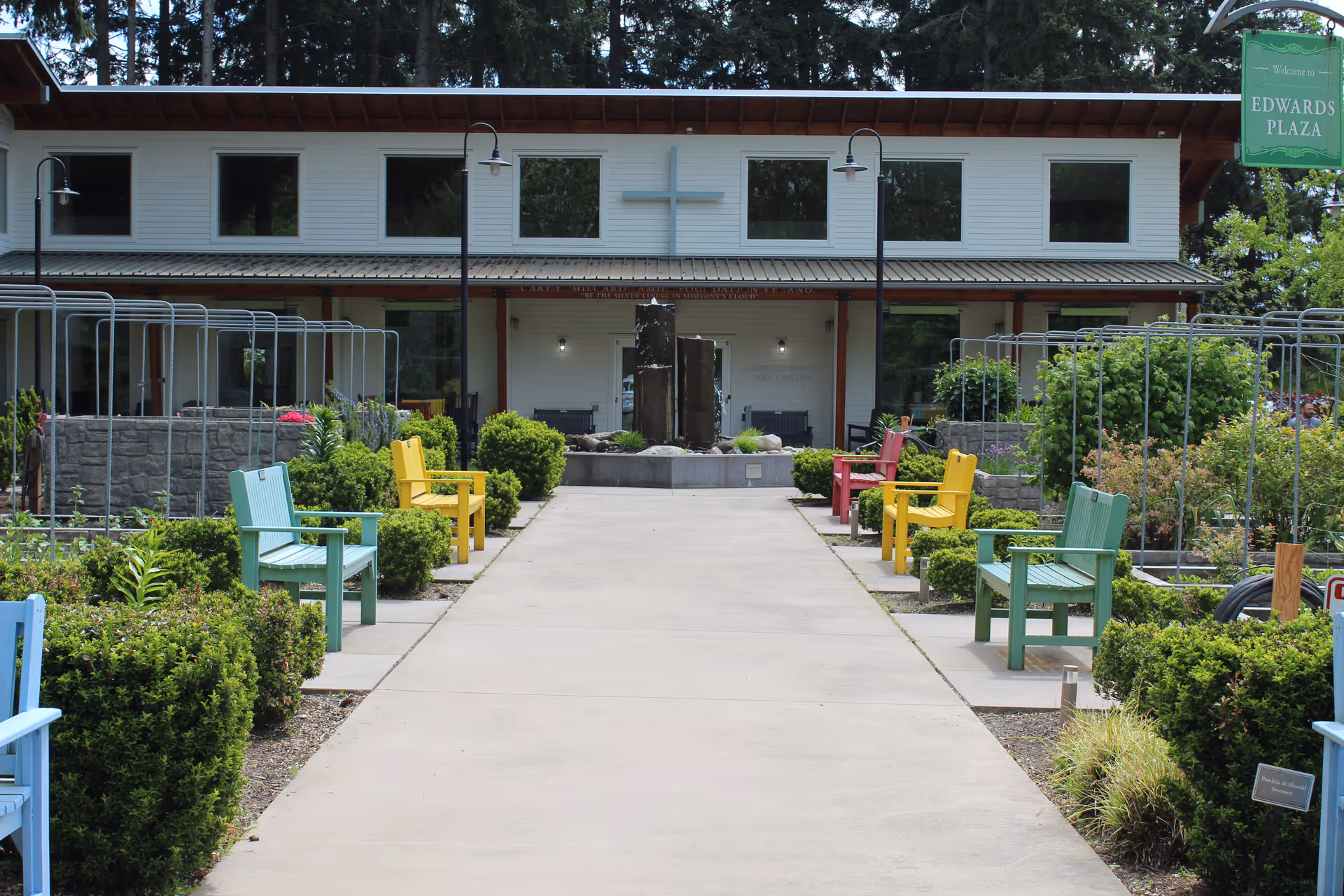 Outdoor garden area with a concrete pathway lined by colorful wooden benches in blue, green, yellow, and red. The pathway leads to a building with large windows and a cross mounted on the wall above the entrance. There are green bushes and plants along both sides of the pathway, and a green sign on the right side reads 'Welcome Edwards Plaza.'
