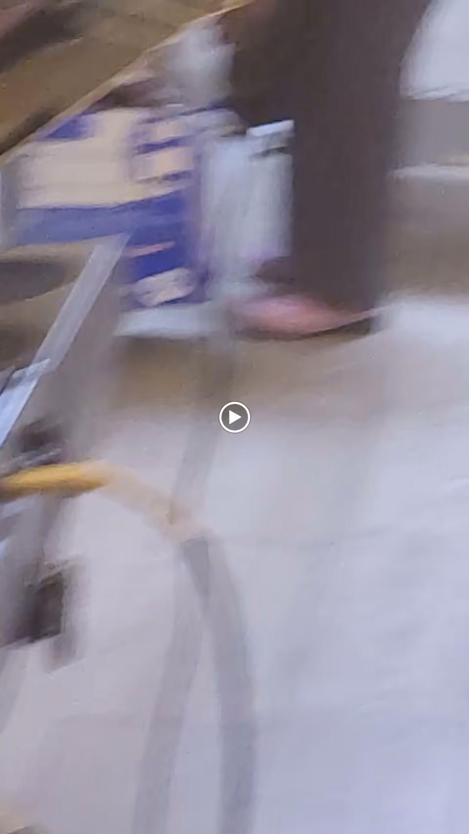 A blurry image showing part of a chair or walker with a yellow handle and a person wearing dark pants and pink slippers standing on a tiled floor.