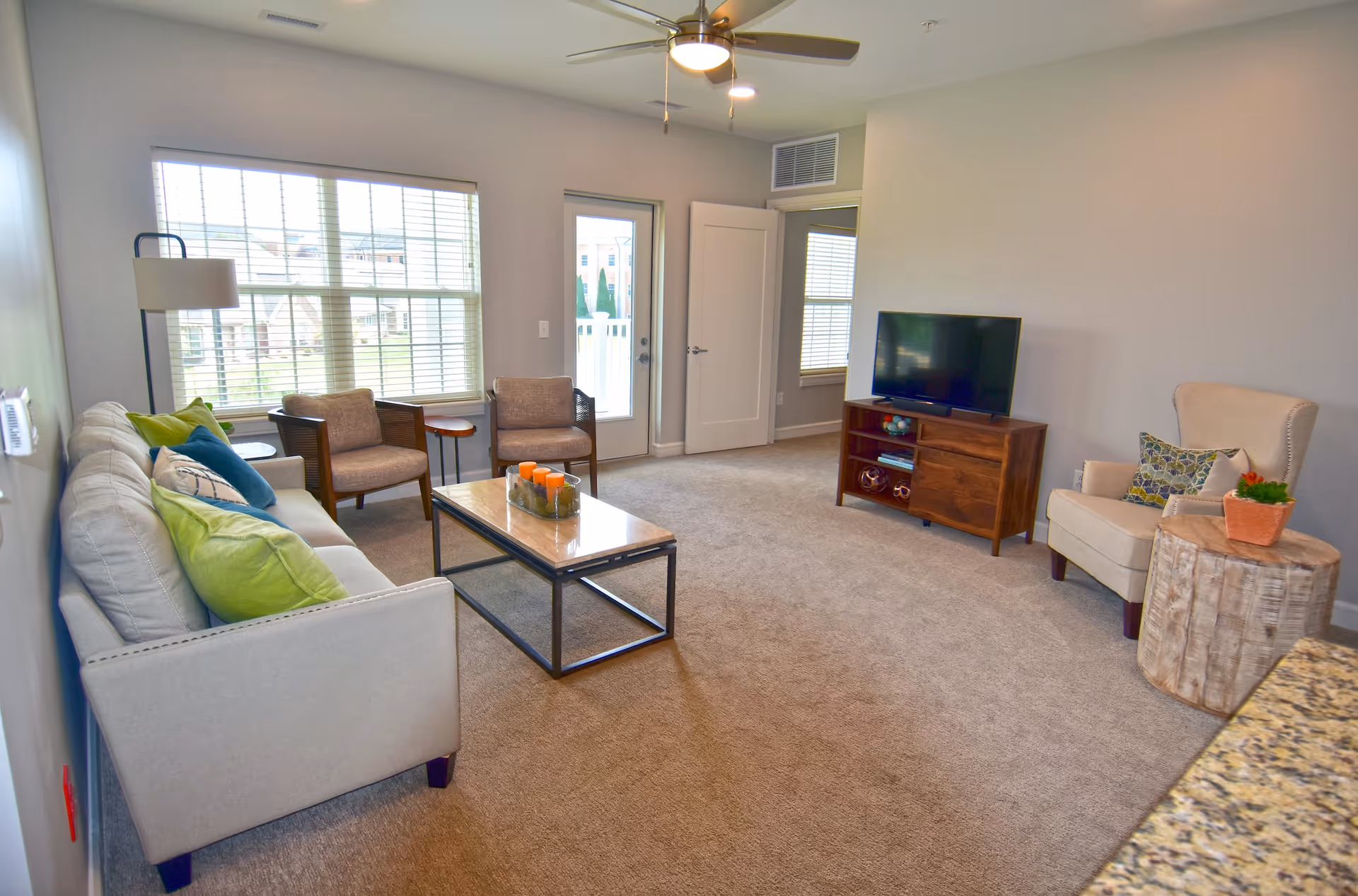 A bright living room with beige carpet and light gray walls. The room features a light gray sofa with colorful pillows, two brown armchairs, a wooden coffee table with orange candles, a beige armchair with patterned pillows, and a wooden TV stand with a flat-screen TV. Large windows with white blinds and a glass door leading outside provide natural light. A ceiling fan with a light is mounted on the ceiling.