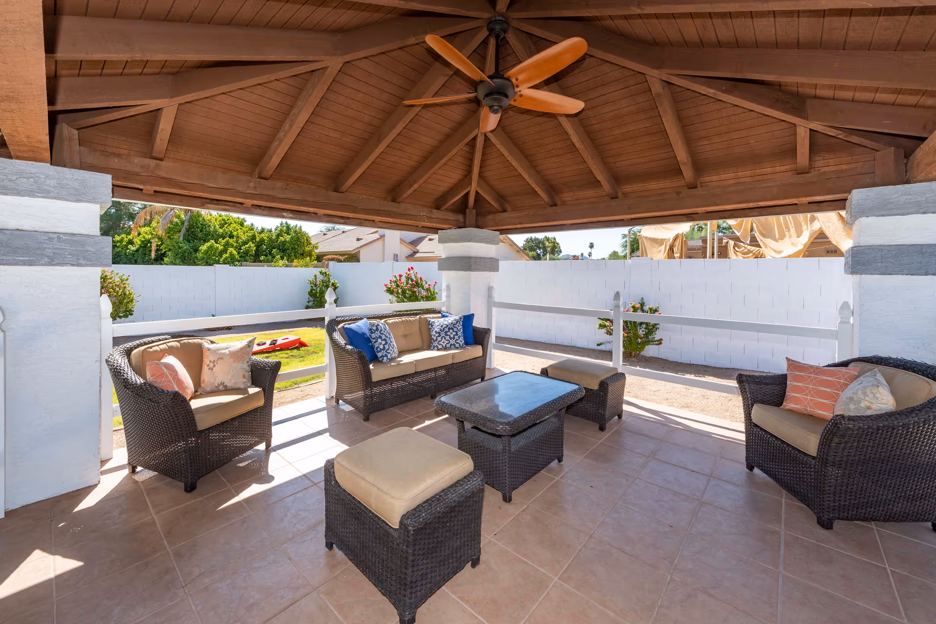 Outdoor covered patio area with a wooden ceiling and ceiling fan, furnished with wicker chairs, a loveseat, cushioned ottomans, and a coffee table. The patio overlooks a garden with green grass, flowering bushes, and a white fence.