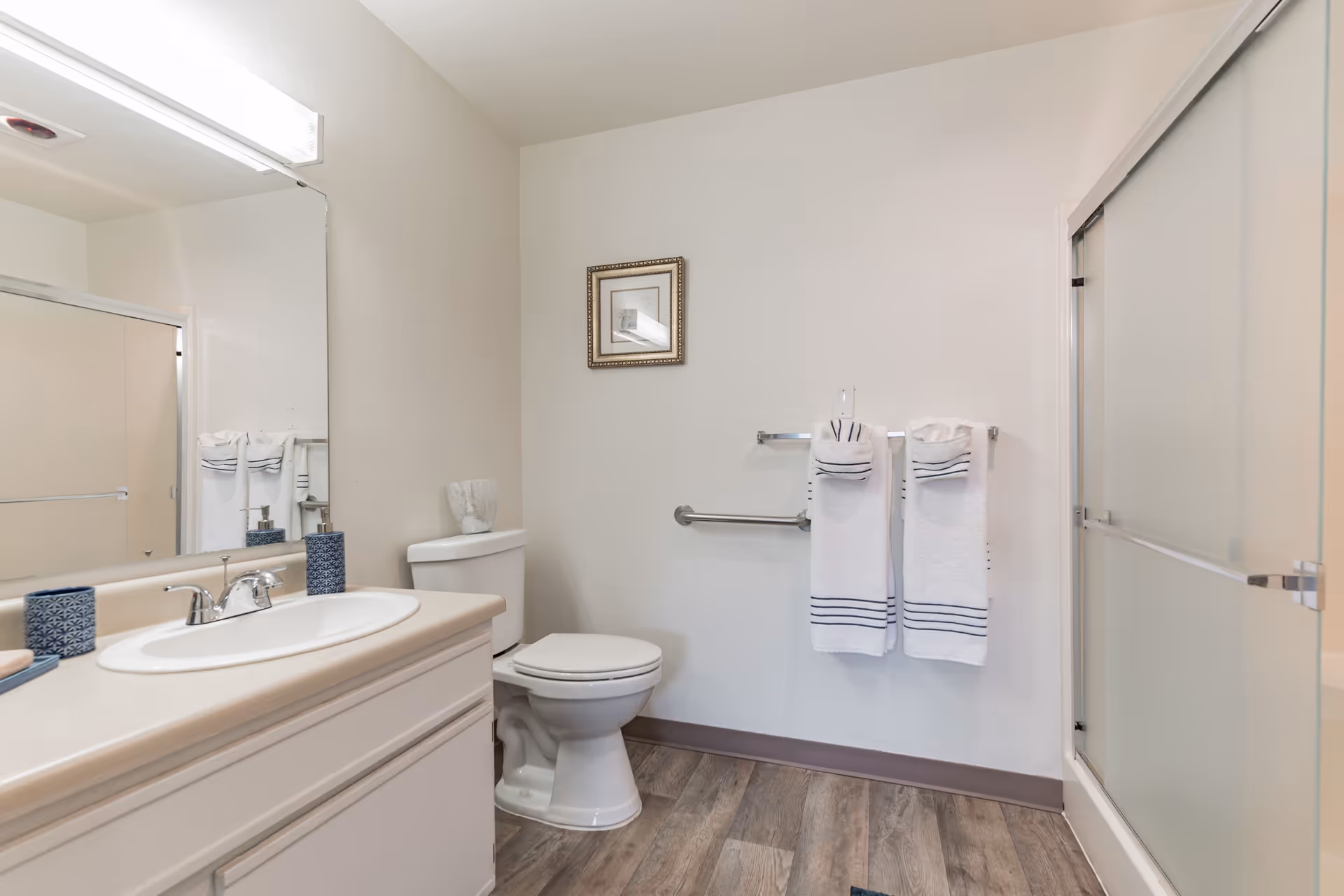 A clean bathroom featuring a white toilet, a vanity with a sink and faucet, a large mirror above the vanity, a shower with frosted sliding doors, and two white towels with blue stripes hanging on a towel rack. There is a small framed picture on the wall above the towels and a decorative item on the toilet tank. The floor has wood-like vinyl planks.