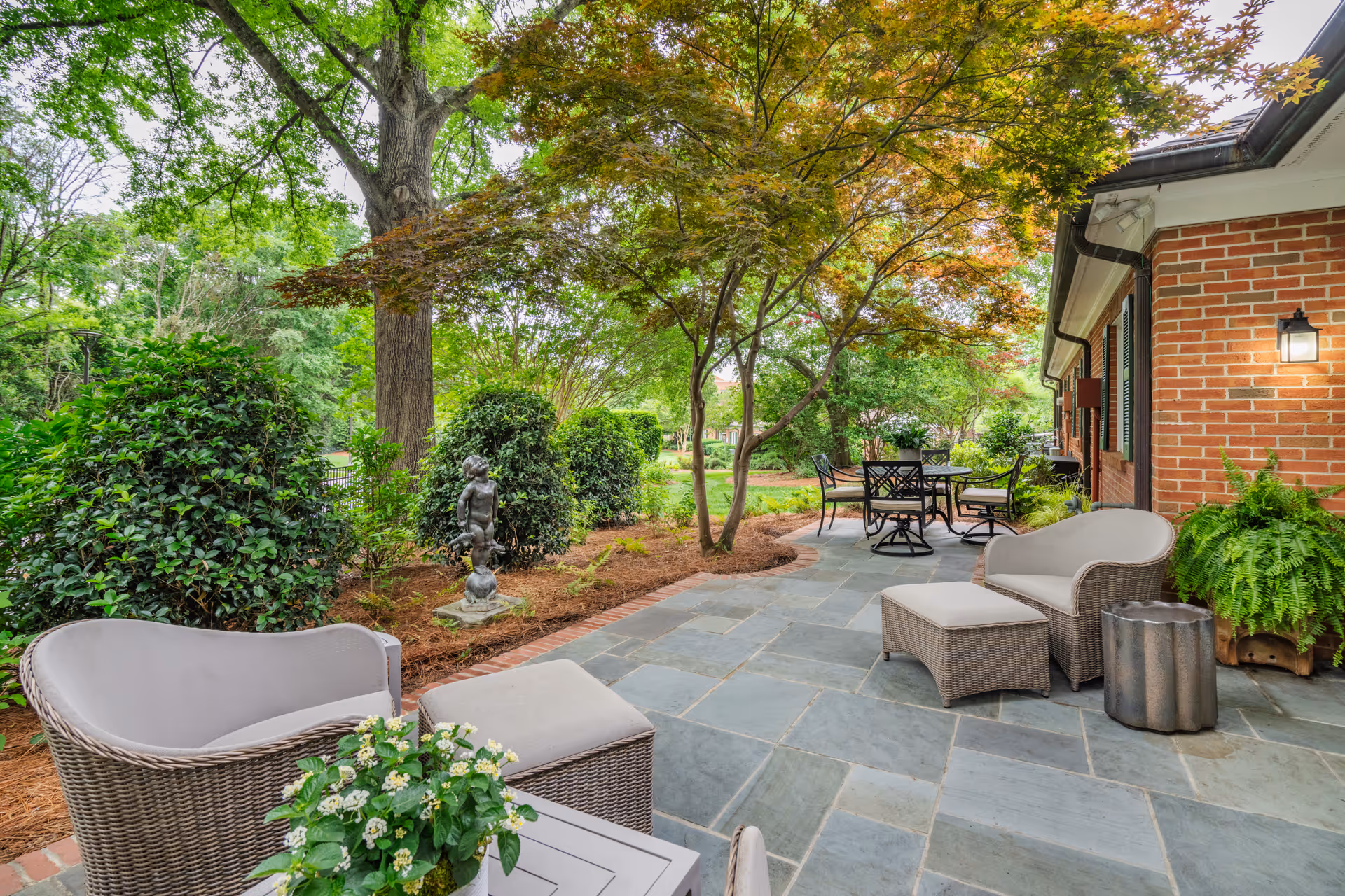 A shaded stone patio with wicker seating, a dining table, and lush landscaping beside a brick building.