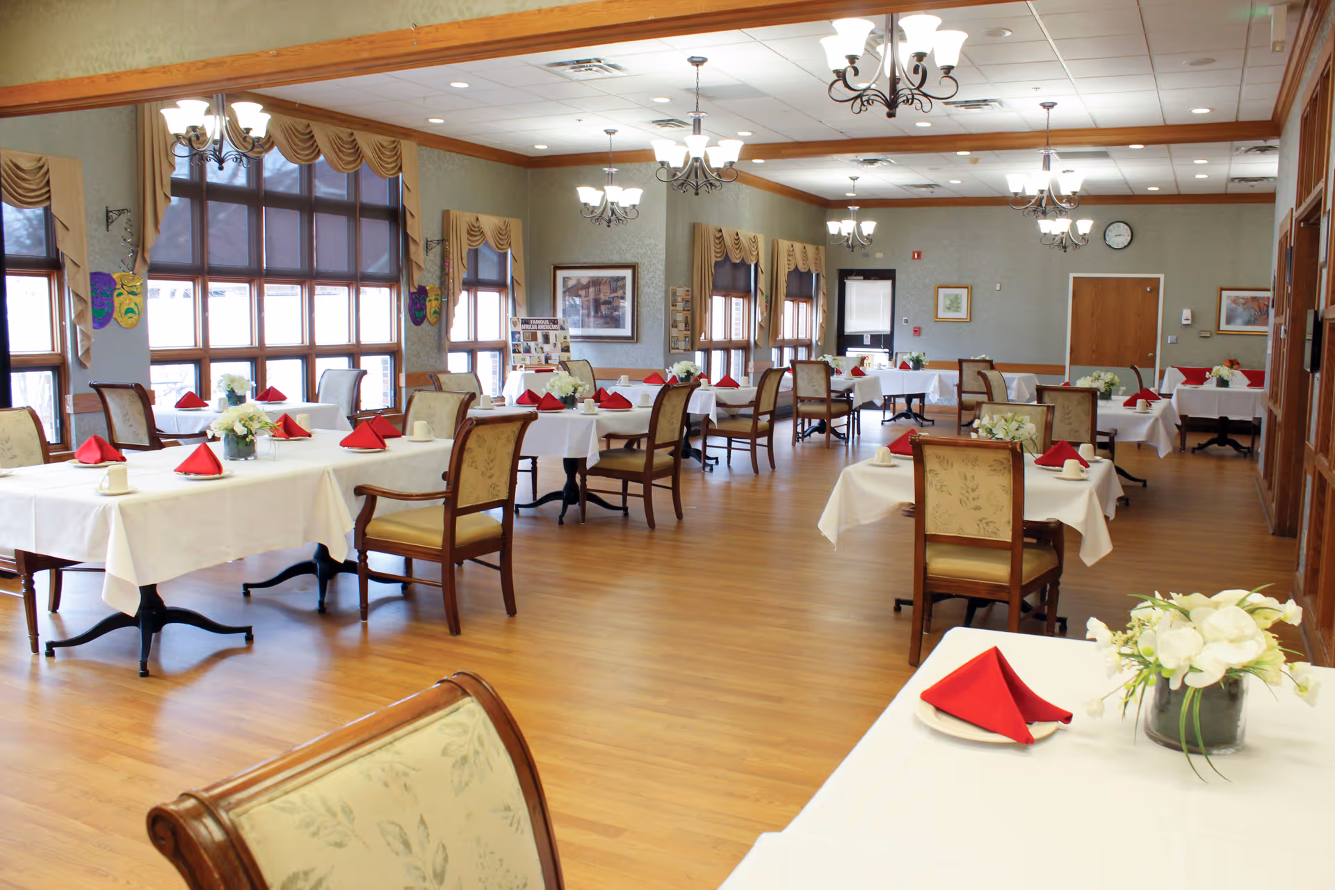 Spacious dining room with neatly set tables, white tablecloths, red napkins, floral centerpieces, and chairs.