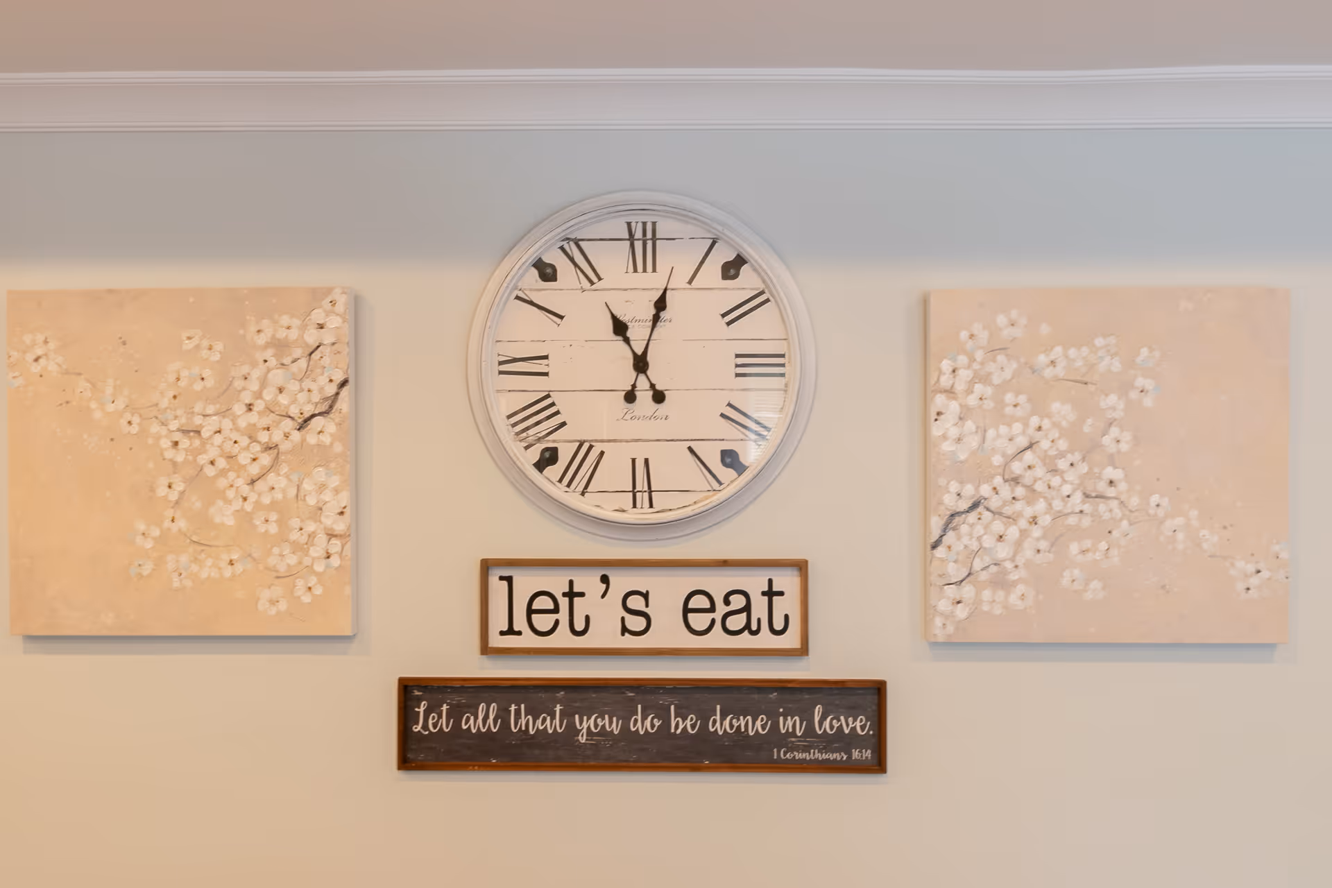 Decorative wall featuring a large round clock between two floral canvases and signs reading "let's eat" and "Let all that you do be done in love."