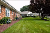 A well-maintained grassy lawn area bordered by a brick building with multiple windows on the left and a large tree on the right, under a cloudy sky.