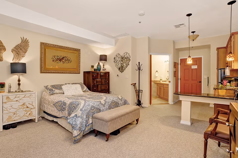 A cozy bedroom with a bed covered in a patterned blue and beige comforter, a beige upholstered bench at the foot of the bed, a wooden dresser, and a nightstand with a lamp. The room has beige walls and carpeted floor. There is a heart-shaped mirror on the wall, a coat rack, and an open doorway leading to a bathroom. To the right, a kitchen area with wooden cabinets and pendant lights is visible.