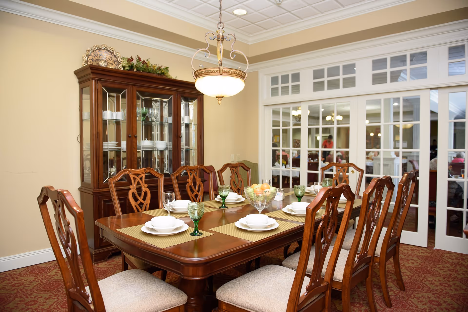 A formal dining room with a large wooden table set for eight people. The table has beige placemats, white dishes, and green glass goblets. Behind the table is a wooden china cabinet with glass doors displaying plates and glassware. The room has beige walls, a red patterned carpet, and a decorative hanging light fixture. In the background, through glass-paneled double doors, another dining area with people is visible.