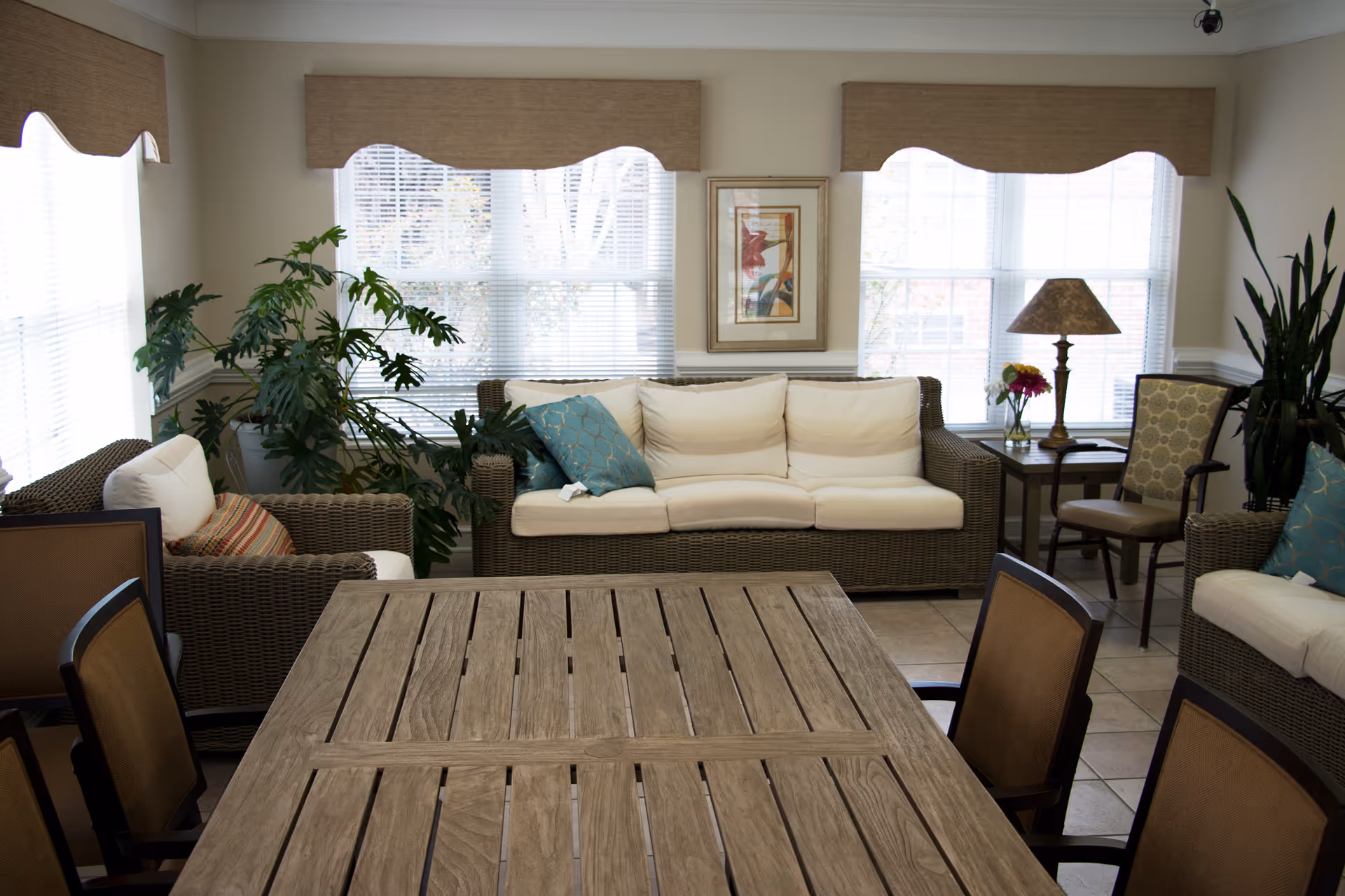 A bright and cozy living room area with a large wooden table in the foreground surrounded by chairs. In the background, there are two wicker sofas with white cushions and decorative pillows, a side table with a lamp and a vase of flowers, framed artwork on the wall, and large windows with blinds letting in natural light. Several green plants add a touch of nature to the room.