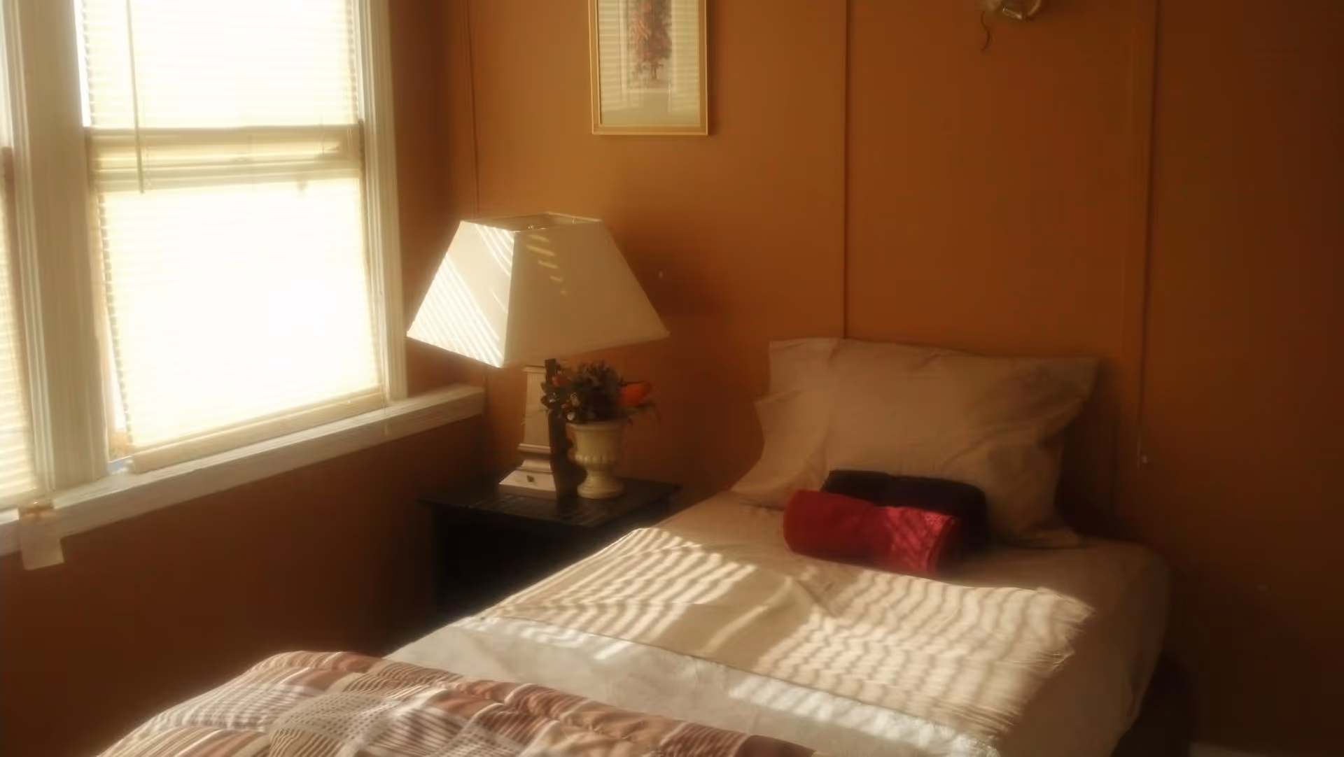Sunlit single bedroom with a bed, nightstand, lamp, and window blinds.