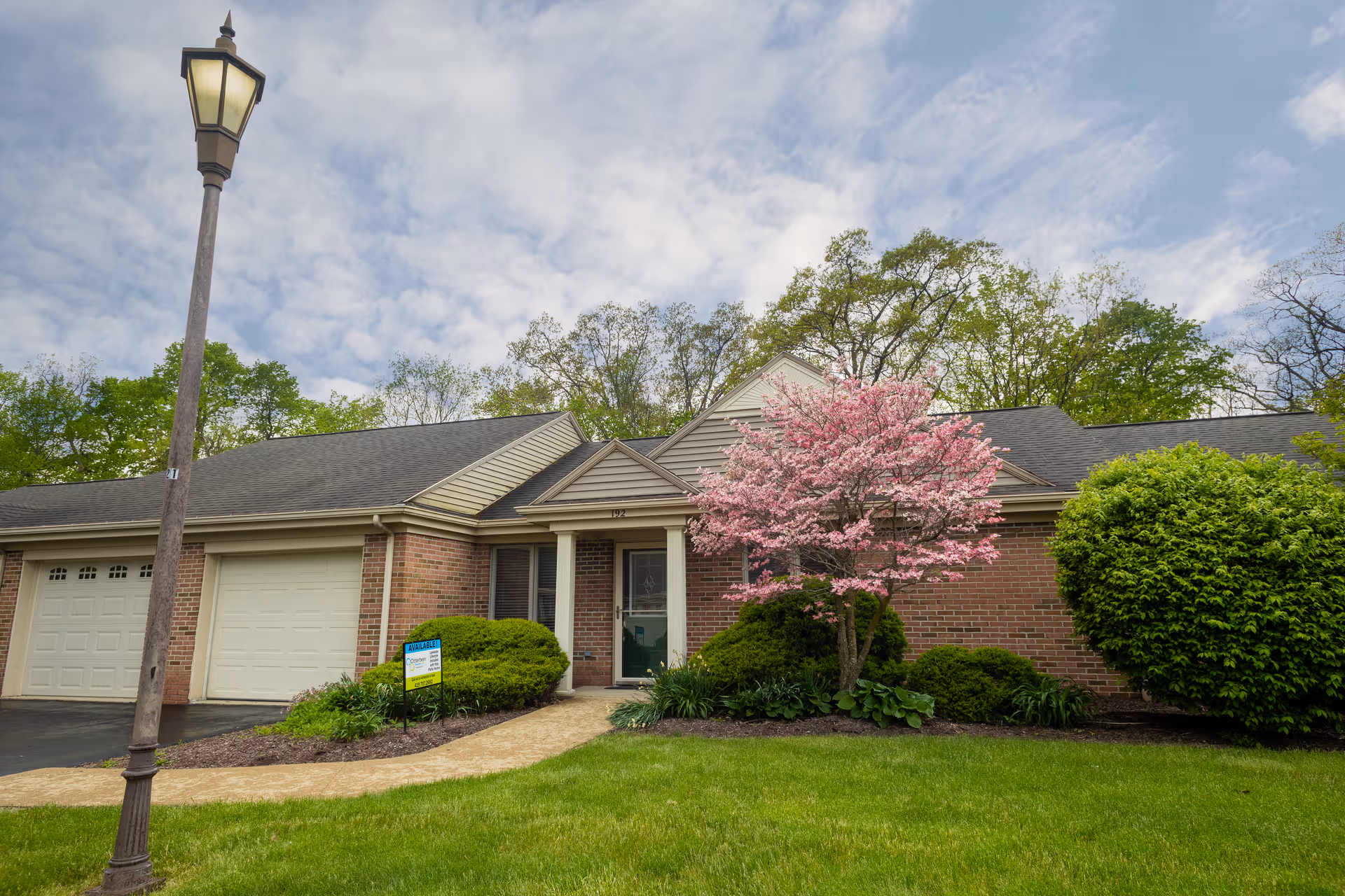 Exterior view of a single-story brick building with a garage, a front door, and a blooming pink tree in the landscaped front yard under a partly cloudy sky.