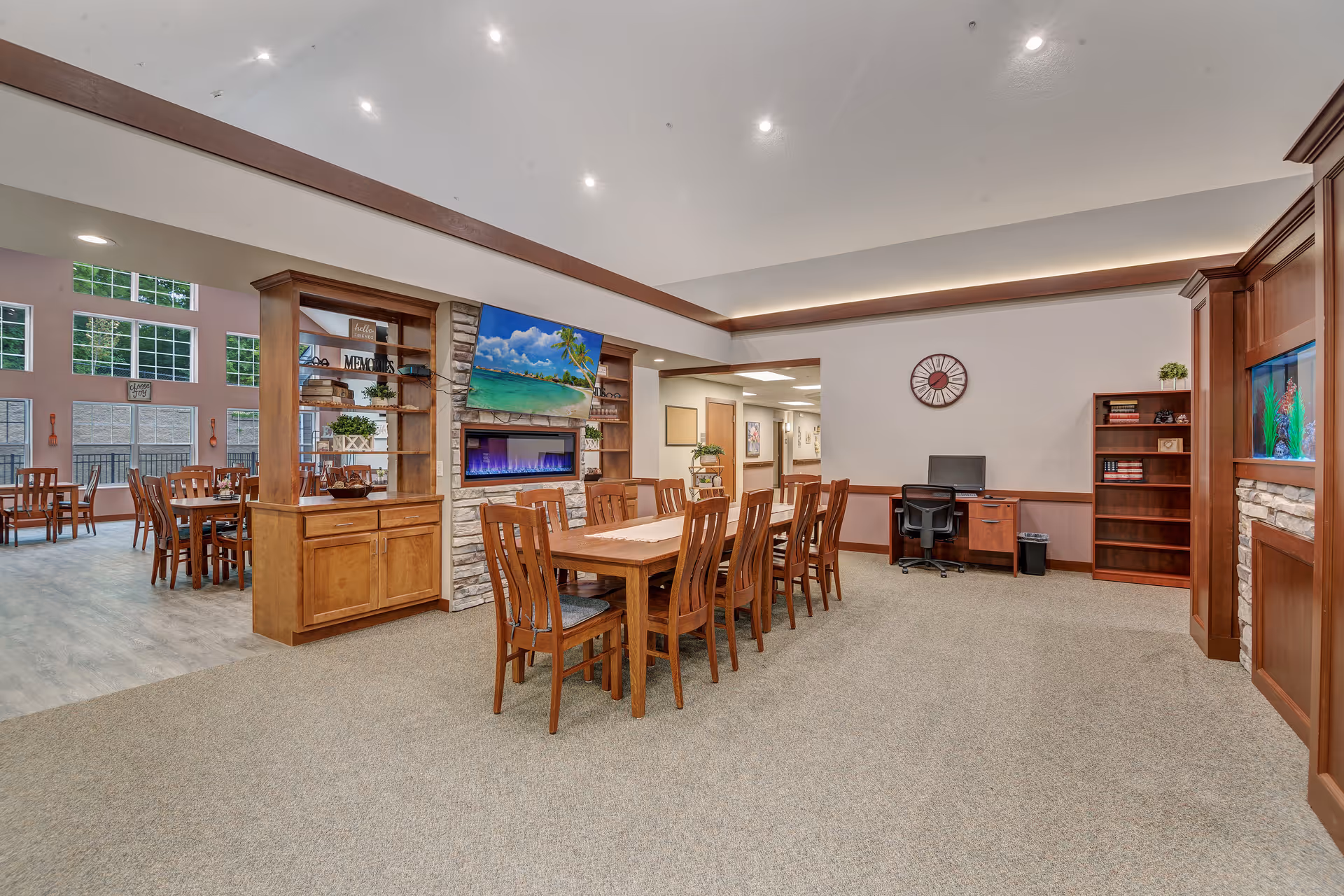 Spacious senior living common area with a long wooden dining table and chairs, built-in shelving and fireplace with TV, and a desk workspace.