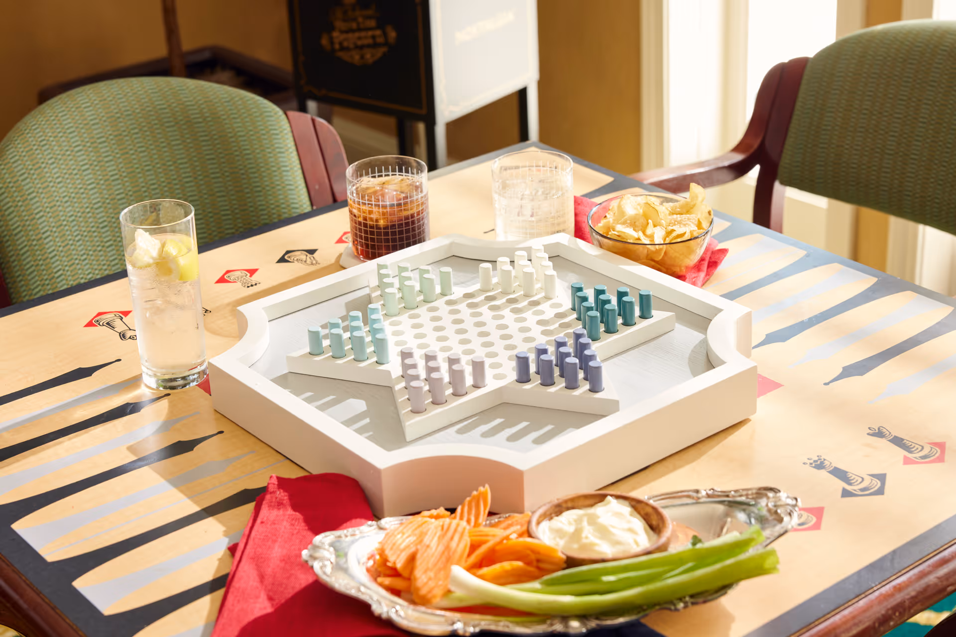 A table set with a Chinese checkers board game, three glasses of beverages including iced tea and water with lemon, a bowl of potato chips, and a silver tray with carrot sticks, celery, and a bowl of dip. The table has a decorative pattern and is surrounded by green cushioned chairs.