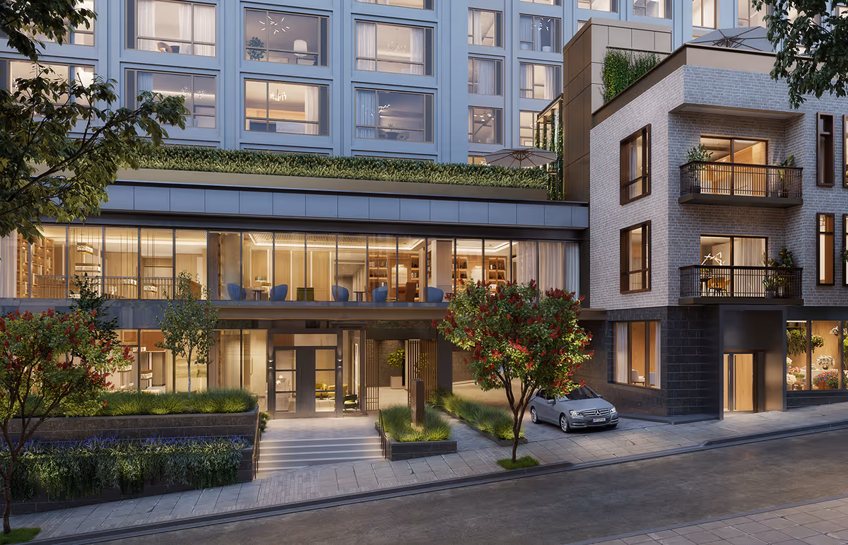 Exterior view of a modern multi-story residential building with large windows, balconies, and a landscaped entrance featuring trees and shrubs. A silver car is parked near the entrance on a paved street.