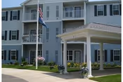 Exterior view of a senior living facility building with white siding, multiple windows, and a covered entrance supported by white columns. There are two flagpoles with flags in front of the building, along with some landscaping including flowers and shrubs.