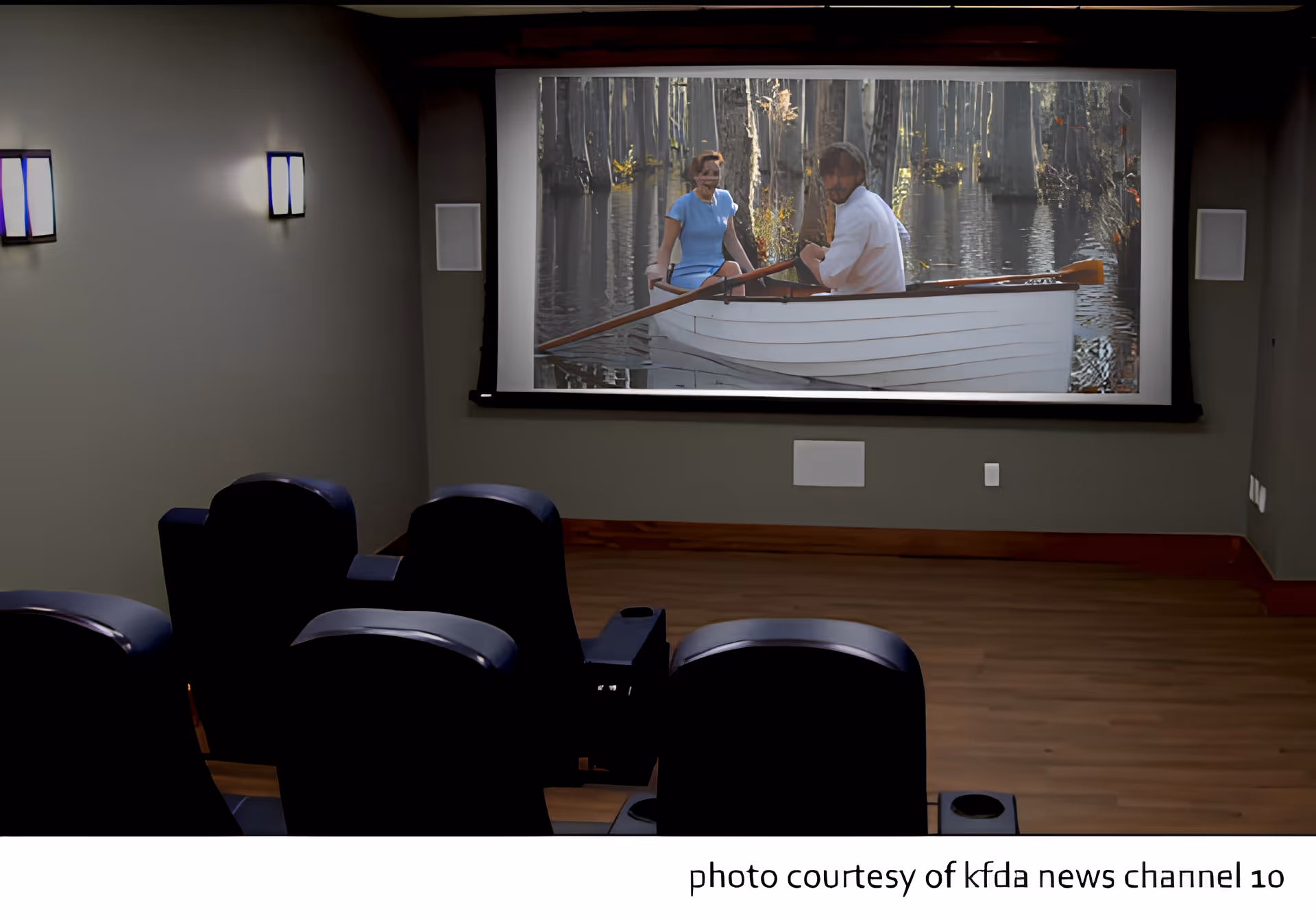 A small home theater room with four black leather recliner chairs facing a large screen showing a movie scene of two people in a white rowboat on a lake surrounded by trees.