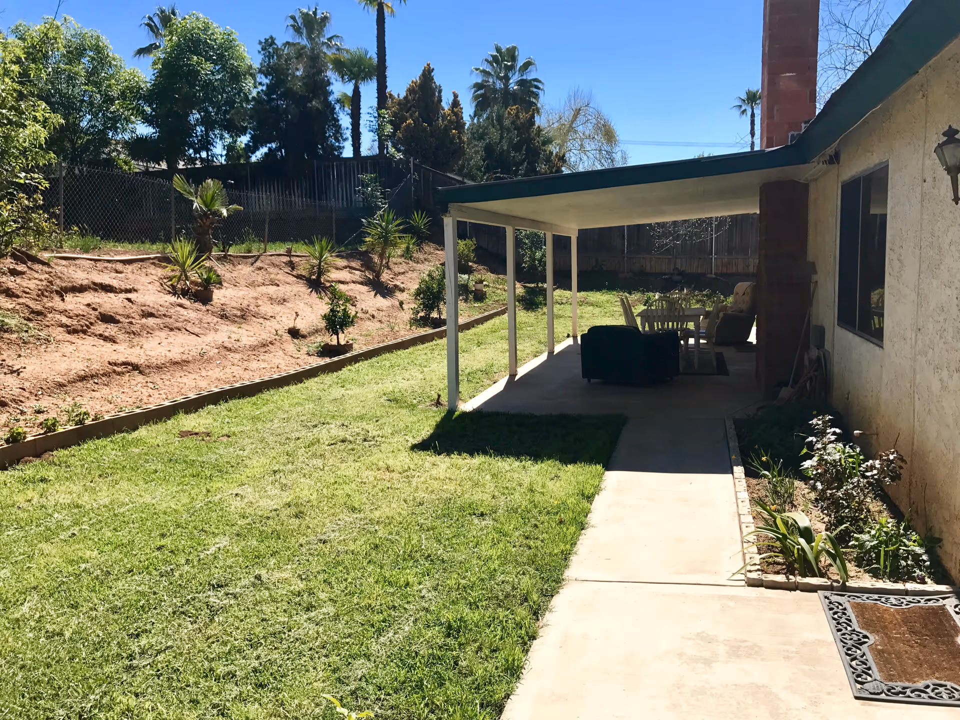 Outdoor view of a residential care facility backyard with a covered patio area featuring outdoor furniture including chairs and a table. The yard has a grassy lawn, a concrete walkway, and some small plants along the edge. Trees and a fence are visible in the background under a clear blue sky.