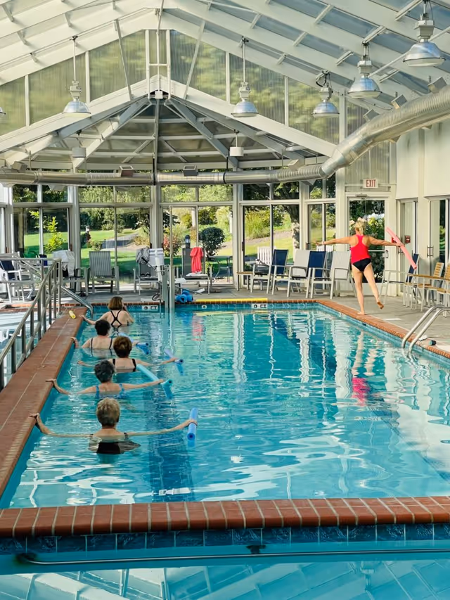 Indoor swimming pool with a group of older adults participating in a water exercise class using pool noodles. The pool area has a glass roof and large windows showing greenery outside. There are chairs and towels along the sides of the pool.