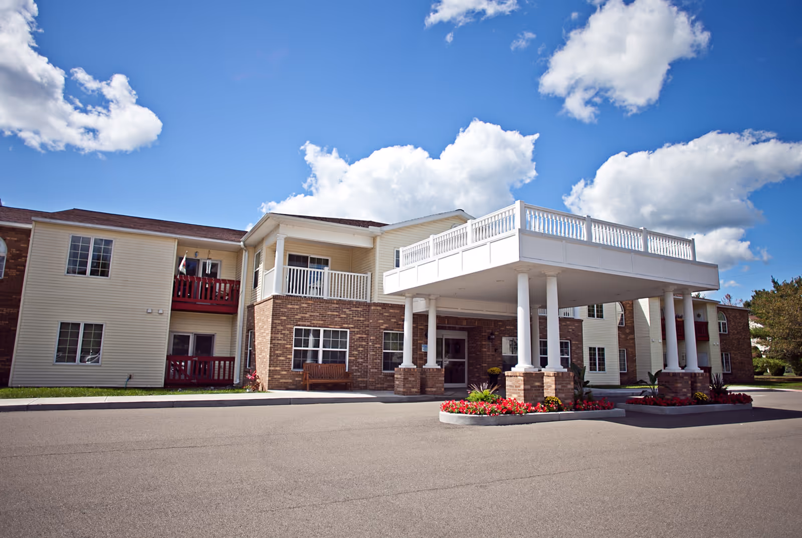 Front entrance of a two-story senior living building with a covered porte-cochere, columns, balconies, and landscaped flower beds under a blue sky.