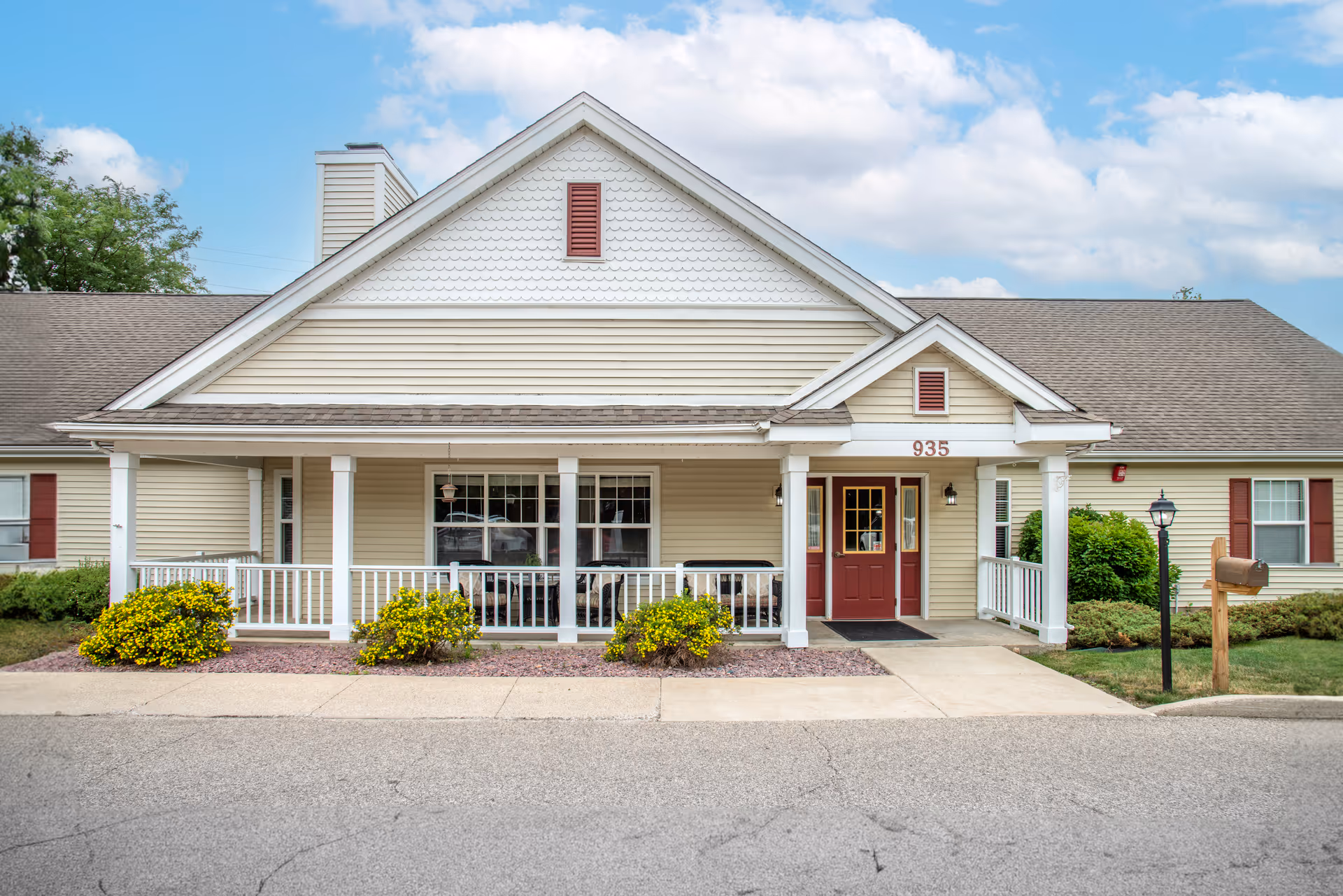 Front exterior view of a single-story building with beige siding and a gabled roof. The building has a covered porch with white railings, red double doors, and the number 935 above the entrance. There are yellow flowering bushes and a mailbox on a wooden post near a black lamp post on the right side. The sky is partly cloudy.