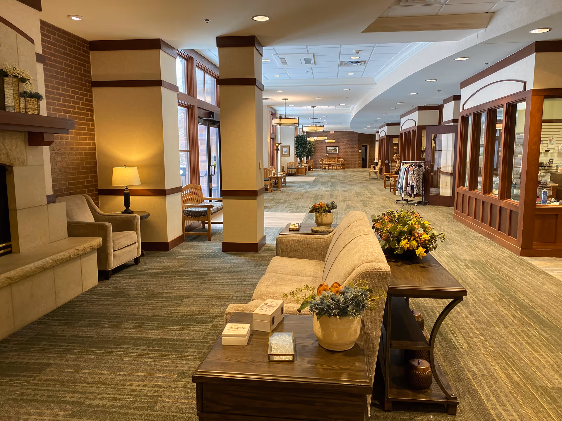 Spacious senior living lobby with sofas, chairs, a fireplace, and a small shop lining a wide carpeted corridor.