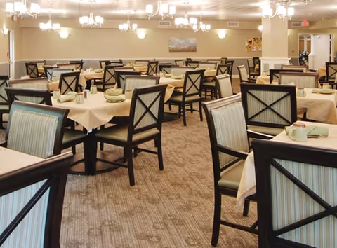 A spacious dining room with multiple tables covered in beige tablecloths, each set with green napkins and cups. The chairs have dark wooden frames with light green striped upholstery. The room is well-lit with ceiling lights and chandeliers, and there are paintings on the walls.