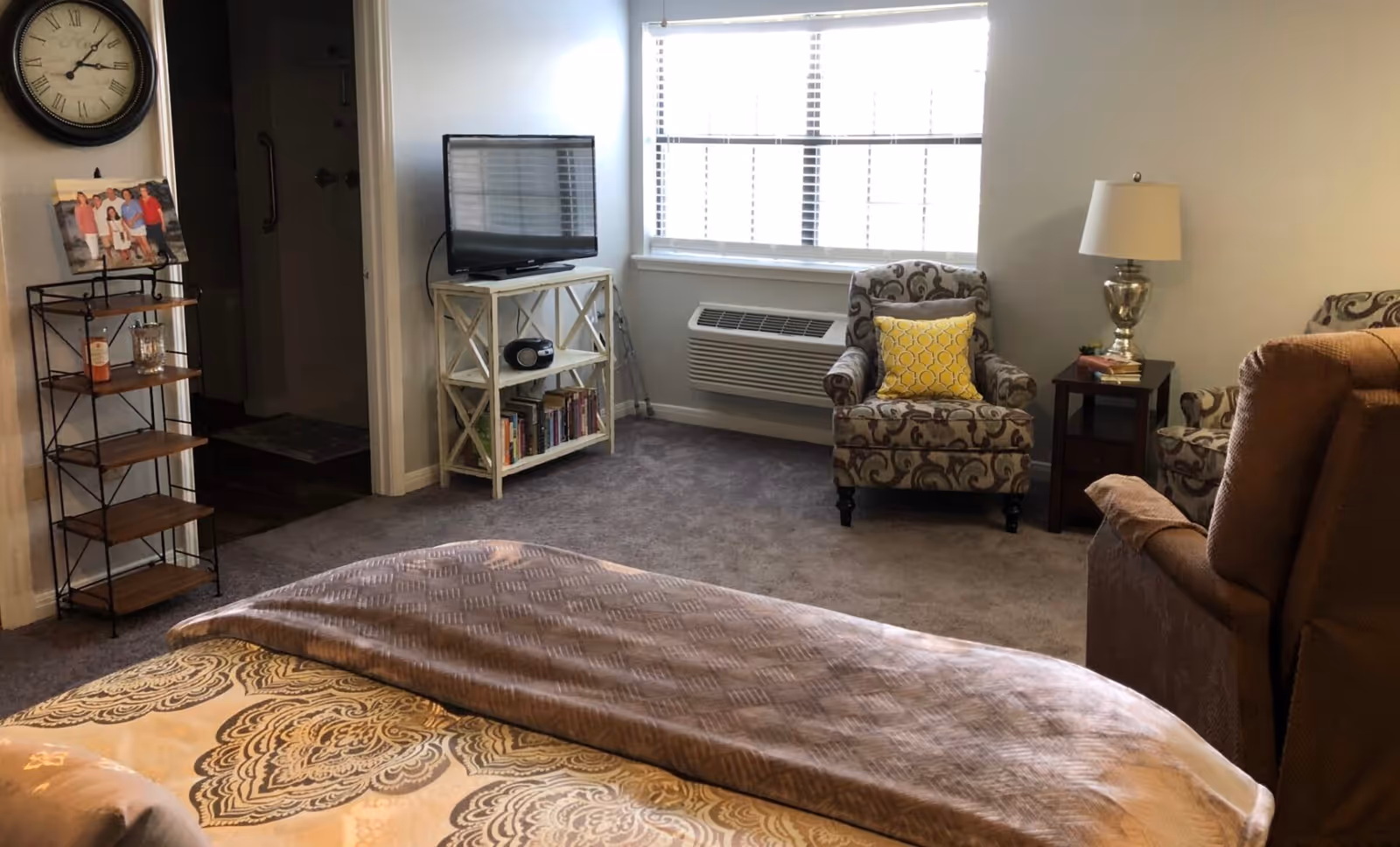 A cozy bedroom with a bed in the foreground covered with a patterned blanket and a brown throw. There is a patterned armchair with a yellow pillow near a window with blinds. A small wooden side table with a lamp and books is next to the armchair. A TV sits on a white shelf unit that also holds books and a radio. A metal and wood shelving unit with a framed photo and a bottle is against the wall near a doorway leading to a bathroom with a visible shower area. A large wall clock is mounted above the shelving unit.
