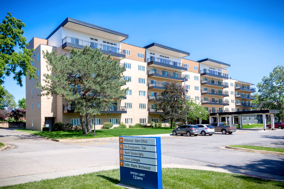 Exterior view of a multi-story senior living facility building with balconies, surrounded by trees and a parking lot with several cars. A blue directional sign in front indicates locations within the facility such as Redwood/Main Office, The Evergreens, North Terrace Apartments, Aspen Cottages, and Cedar, Maple, Birchwood. The sky is clear and blue.