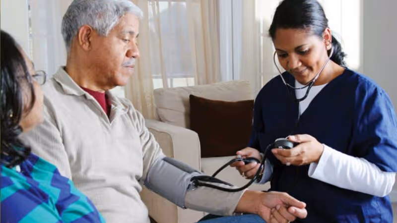 A healthcare professional in navy blue scrubs is measuring the blood pressure of an elderly man seated on a couch in a living room setting, while another person sits nearby.
