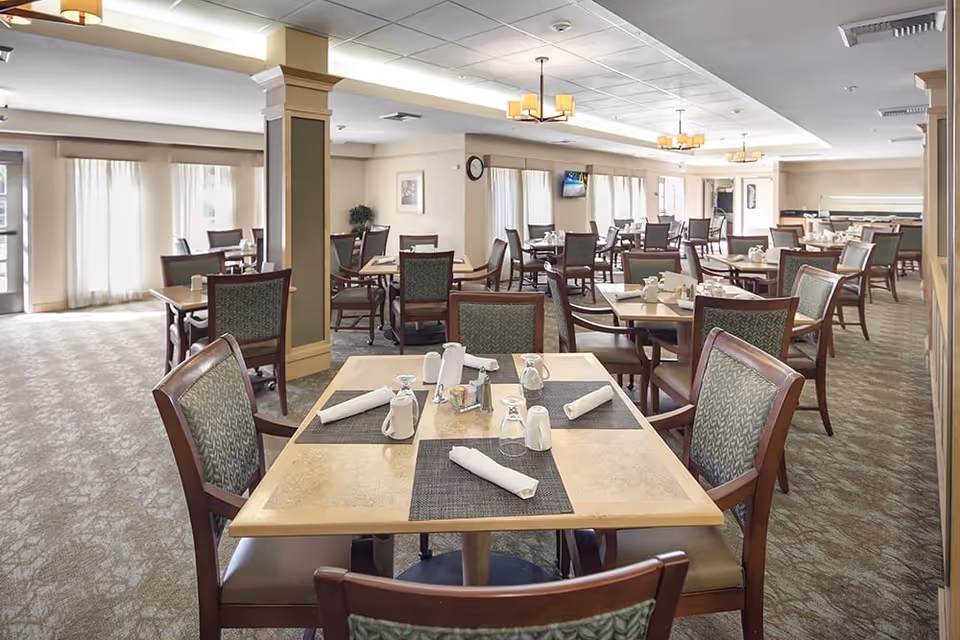 A spacious dining room in a senior living facility with multiple tables and chairs arranged neatly. Each table is set with placemats, napkins, glasses, and condiments. The room has large windows with sheer curtains allowing natural light to fill the space. The ceiling features recessed lighting and hanging light fixtures, and there is a wall clock and a mounted television visible in the background.