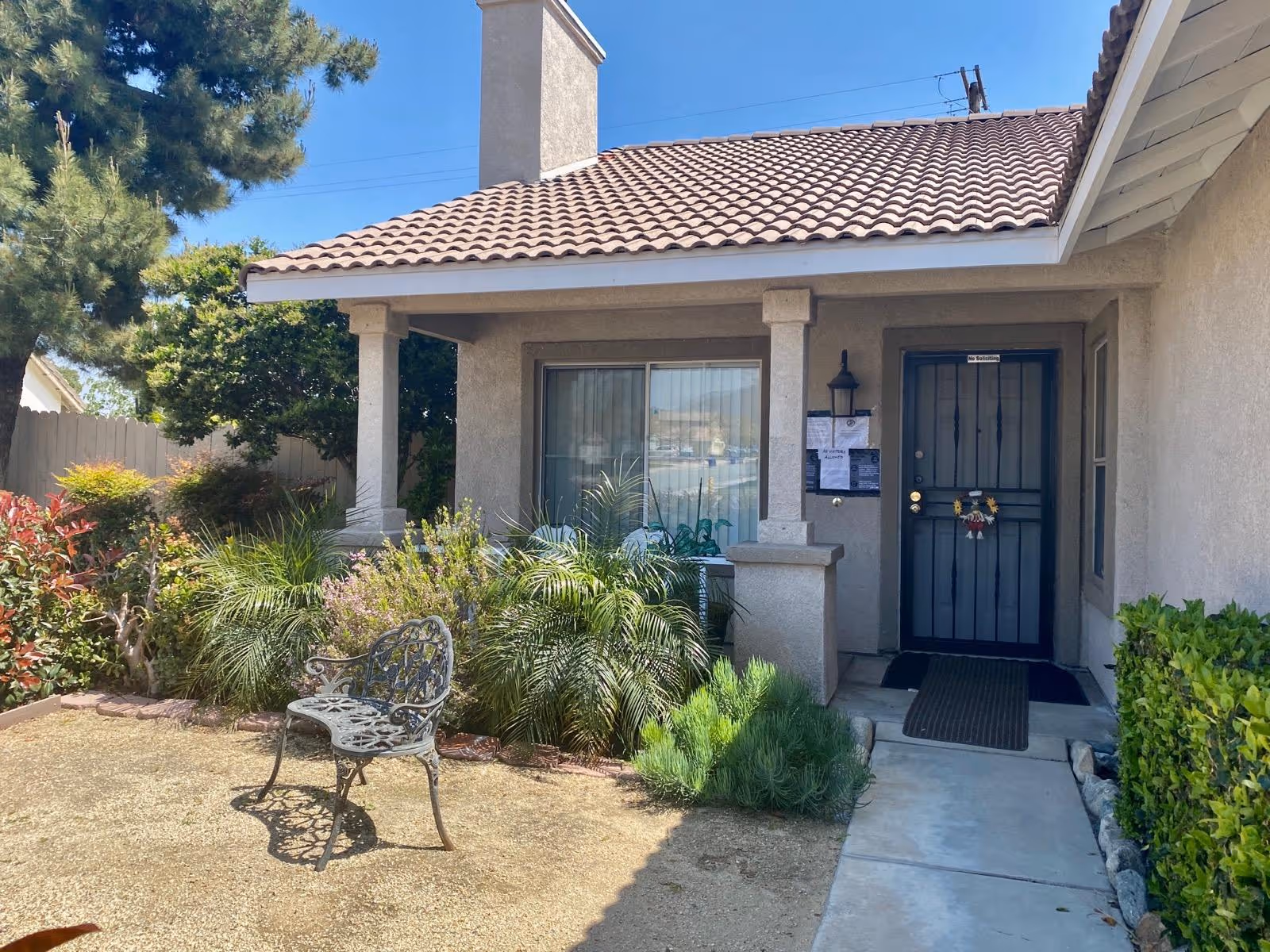 Front exterior view of a single-story house with a tiled roof, a small porch supported by two columns, a black metal front door with a 'No Soliciting' sign, and a window with vertical blinds. The front yard features various green plants, bushes, a tree, and a decorative metal bench on a dirt patch.