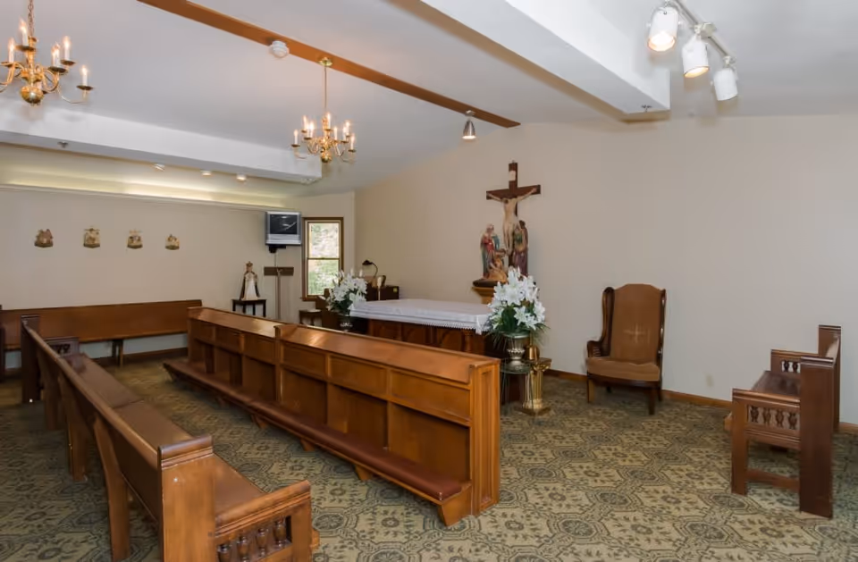 Interior view of a small chapel with wooden pews, an altar adorned with white flowers, a crucifix on the wall, religious statues, chandeliers, and a patterned carpeted floor.