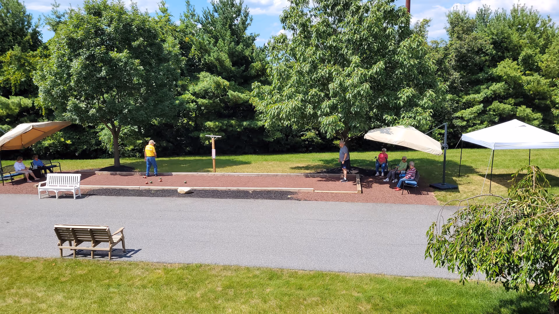 Outdoor scene at Traditions of Hanover showing a bocce ball court with two people playing and several others sitting on benches under umbrellas and trees, surrounded by green grass and trees under a partly cloudy sky.