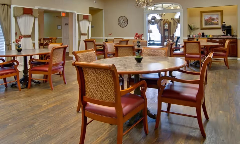 Spacious dining room featuring round wooden tables, upholstered chairs, wood flooring, and a serving counter in the background.