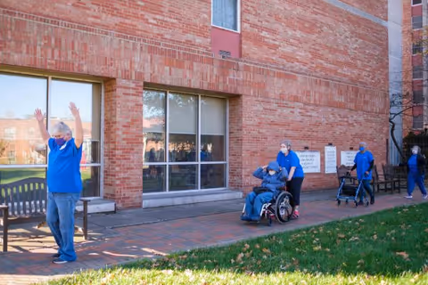 A group of people outside a brick building, with one person in a wheelchair being pushed by a caregiver, another person walking with a walker, and another person raising their arms. All individuals are wearing blue shirts and face masks.