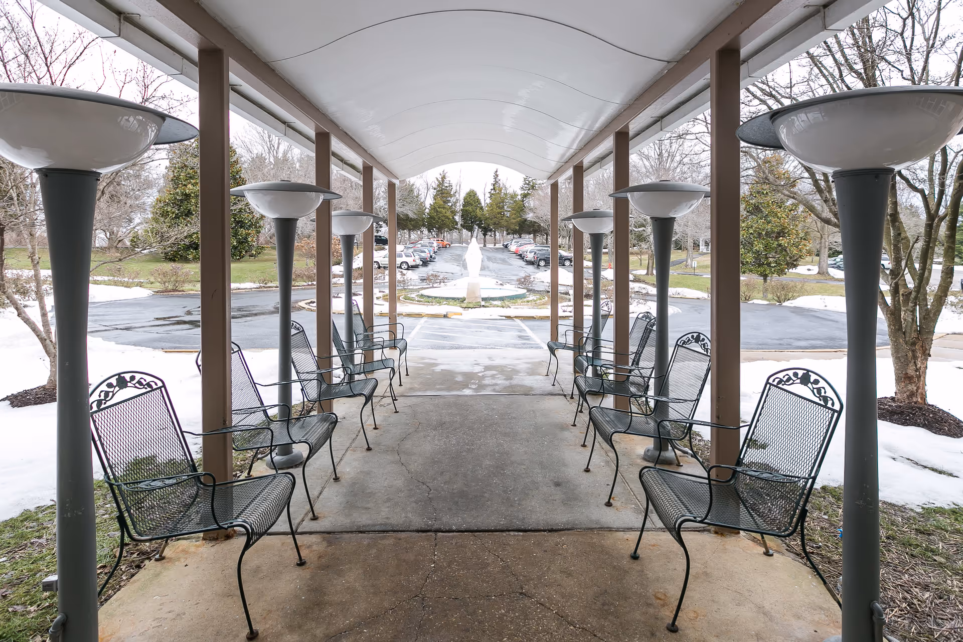 Covered outdoor walkway with metal chairs lined up on both sides and tall lamp posts, leading to a parking lot with cars and trees in the background. Snow is visible on the ground outside the walkway.