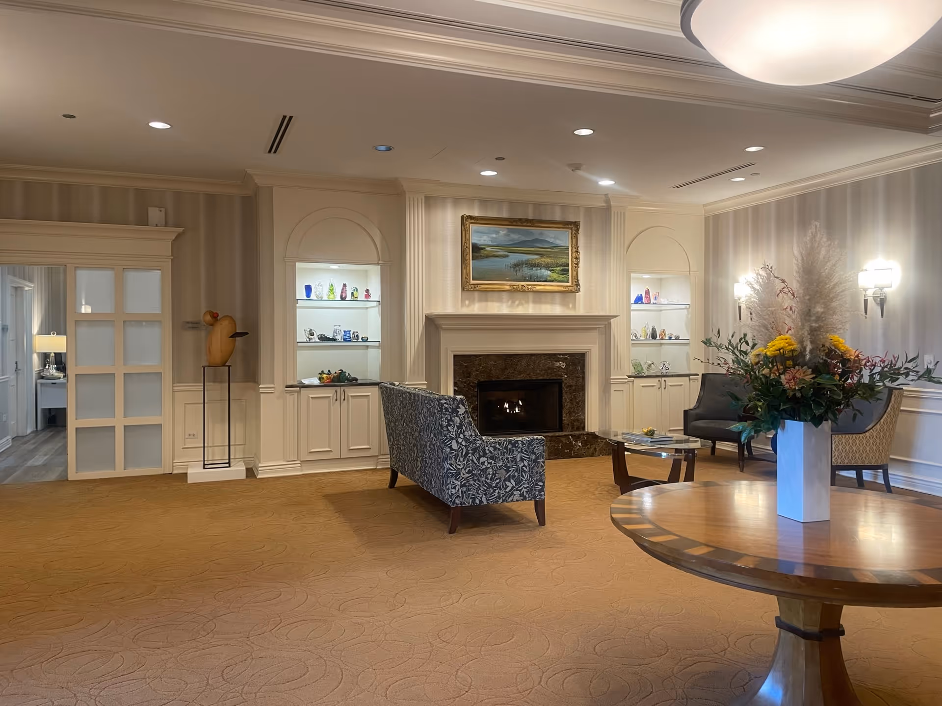 Elegant lounge area with a fireplace, seating, display shelves, and a round table topped with a floral arrangement.