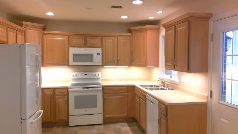 A clean, well-lit kitchen with light wood cabinets, a white refrigerator, a white stove with an overhead microwave, a dishwasher, a double sink, and a door with glass panes leading outside.
