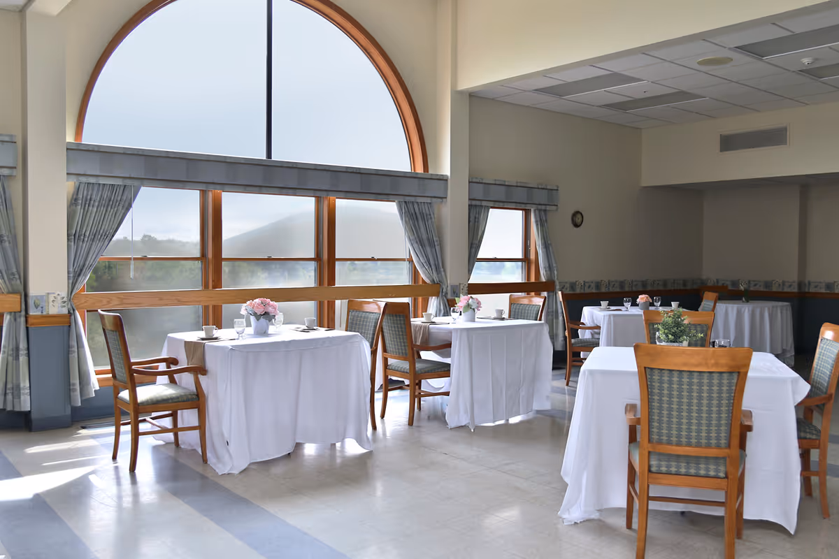 Bright dining room with white-clothed tables and wooden chairs arranged near a large arched window.