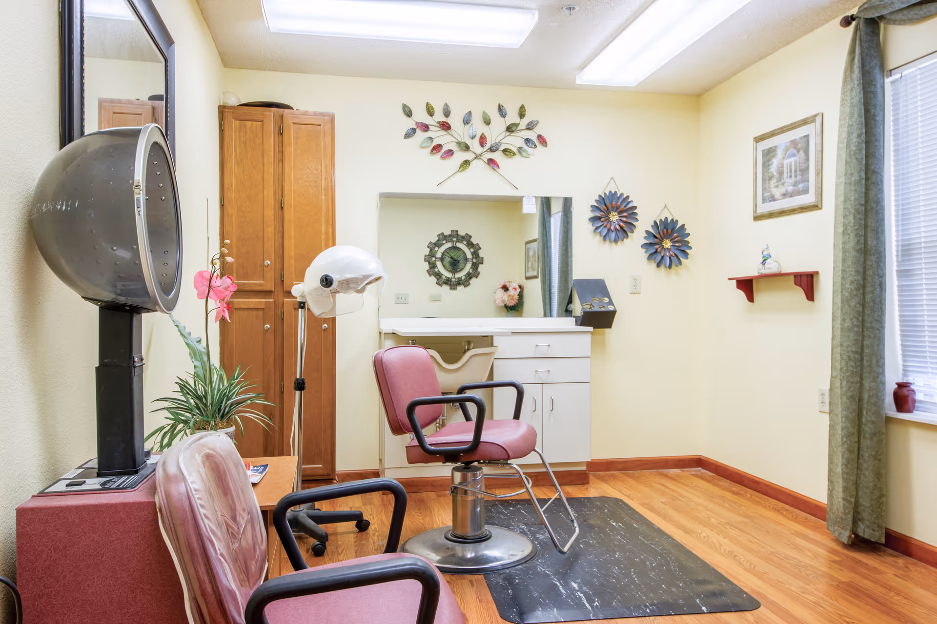 Interior of a small salon room with two maroon salon chairs, a hair dryer hood, a white hair steamer, a wooden cabinet, a large mirror on the wall, decorative wall art, and a window with green curtains letting in natural light.
